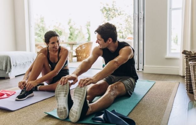 a couple are stretching in a room on the carpet with a sofa and baskets surrounded | Exercise and Immune Function | SARS-CoV-2 and The Implications of Lifestyle and Aging